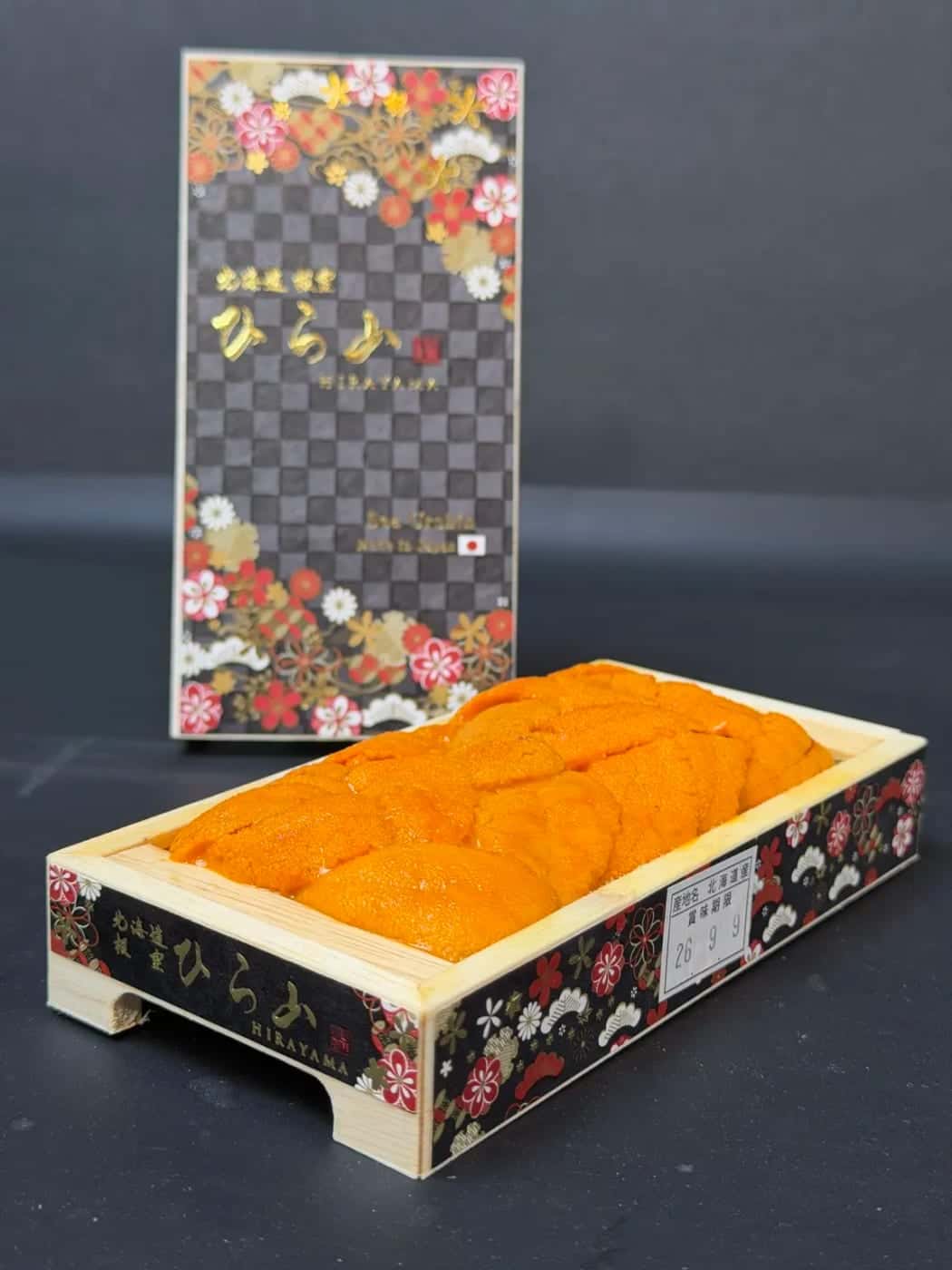 A wooden Hirayama tray of Hokkaido bara uni, lobes bright orange, with its printed sleeve behind