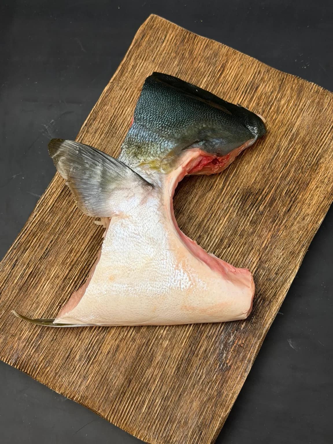 An uncooked Japanese hamachi collar with skin and fin attached on a wooden board.