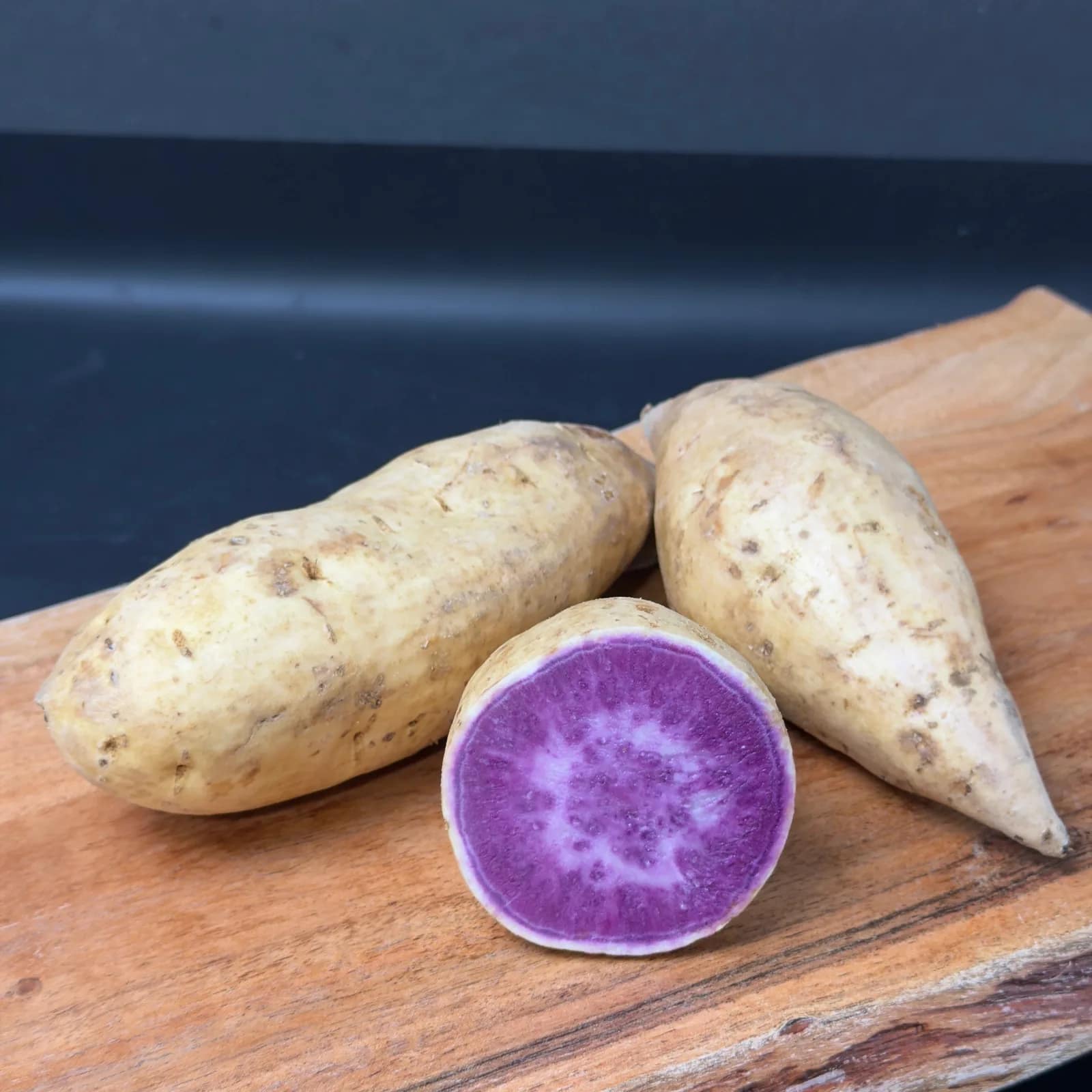 Two whole Okinawan purple sweet potatoes beside a cut half showing purple flesh on a wooden board.