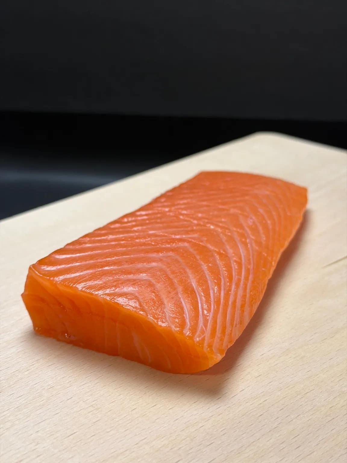 Thawed salmon saku block on a pale wooden board, showing its marbling and cut end.